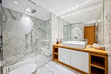 Spacious, modern bathroom featuring white marble walls and floor, a glass shower, toilet, wooden vanity with a vessel sink, and a large mirror.