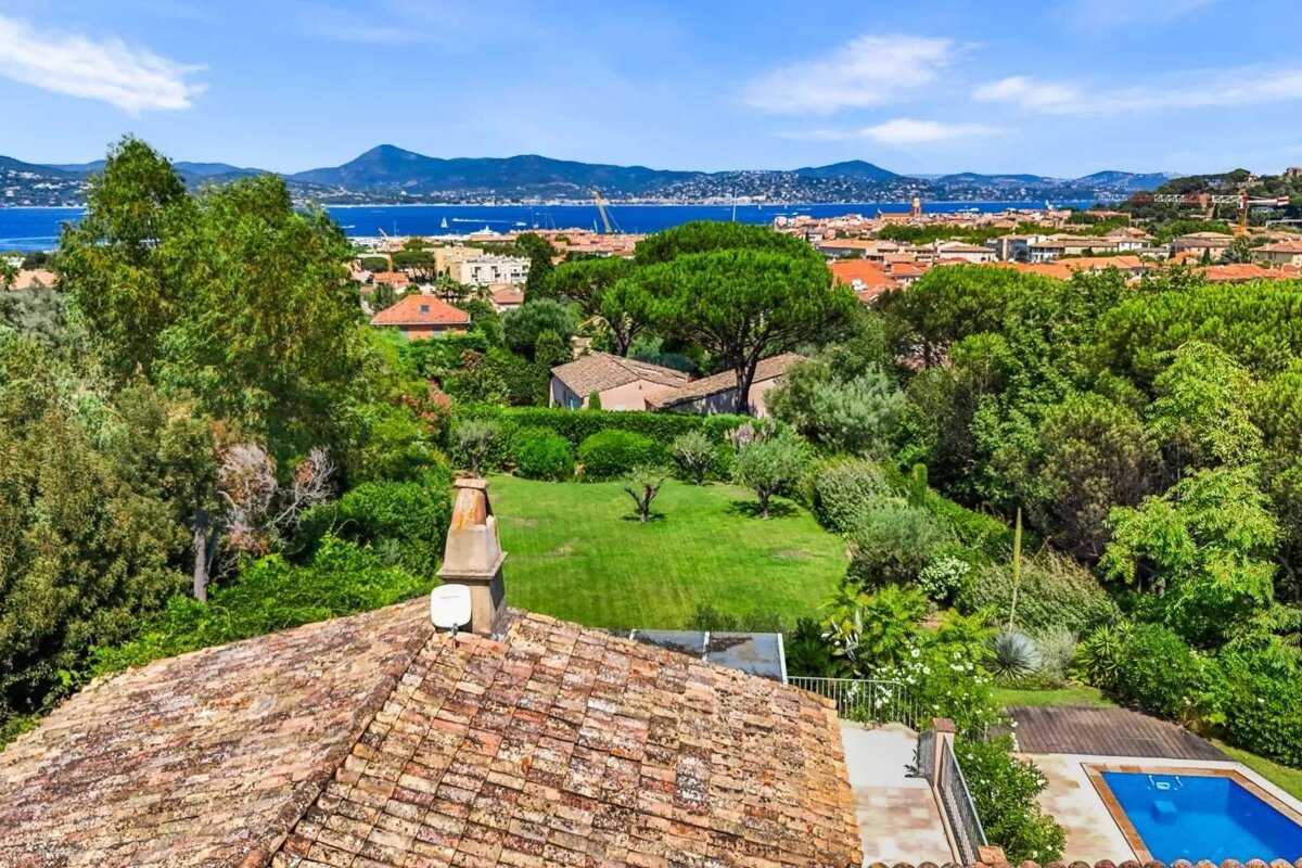 Elevated view of a verdant landscape with red-tiled roofs, a distant blue bay, mountains, and a visible swimming pool.