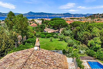 Elevated view of a verdant landscape with red-tiled roofs, a distant blue bay, mountains, and a visible swimming pool.