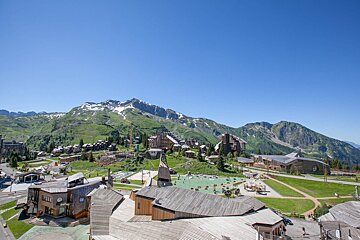 A high-angle view of a modern mountain village with unique wooden buildings, surrounded by green, snow-capped peaks under a clear blue sky.