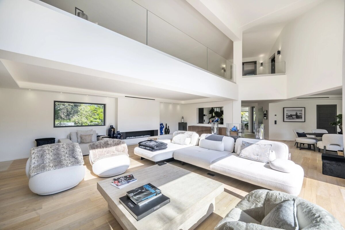 Bright, modern, open-plan living room with high ceilings, white walls, light wood floors, and minimalist white furniture. A balcony overlooks the space.