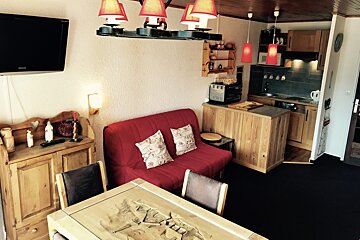 A cozy studio apartment with a red couch, wooden table and kitchen island, a wall-mounted TV, and rustic red light fixtures.