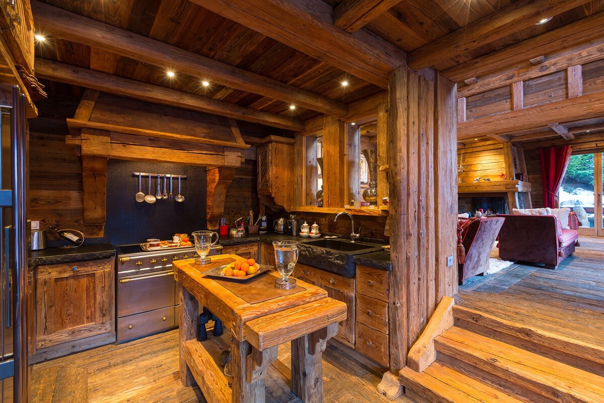 Rustic chalet interior with a wooden kitchen featuring a stove and sink, opening to a cozy living room with a fireplace.