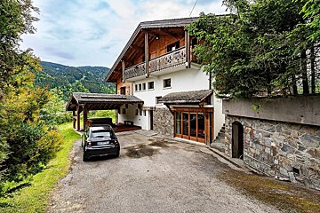 A large, white and wooden chalet stands in a lush, mountainous setting with a black car and covered hot tub in the driveway.
