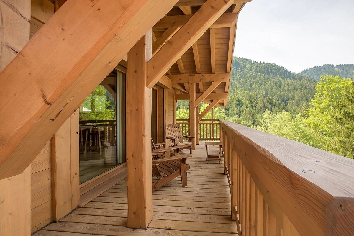A rustic wooden balcony with chairs and a small table overlooks a lush green mountain forest under a bright sky, part of a timber cabin.
