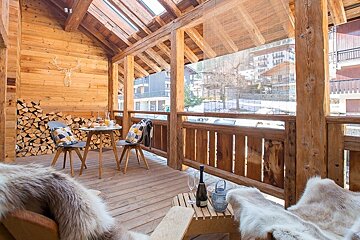 A cozy wooden balcony of a mountain chalet with fur throws, a small dining set, stacked logs, and scenic winter views.