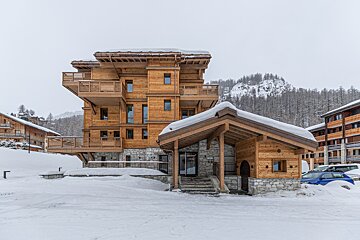 A multi-story wooden ski resort building covered in snow, surrounded by a winter landscape with snow-covered trees and mountains.