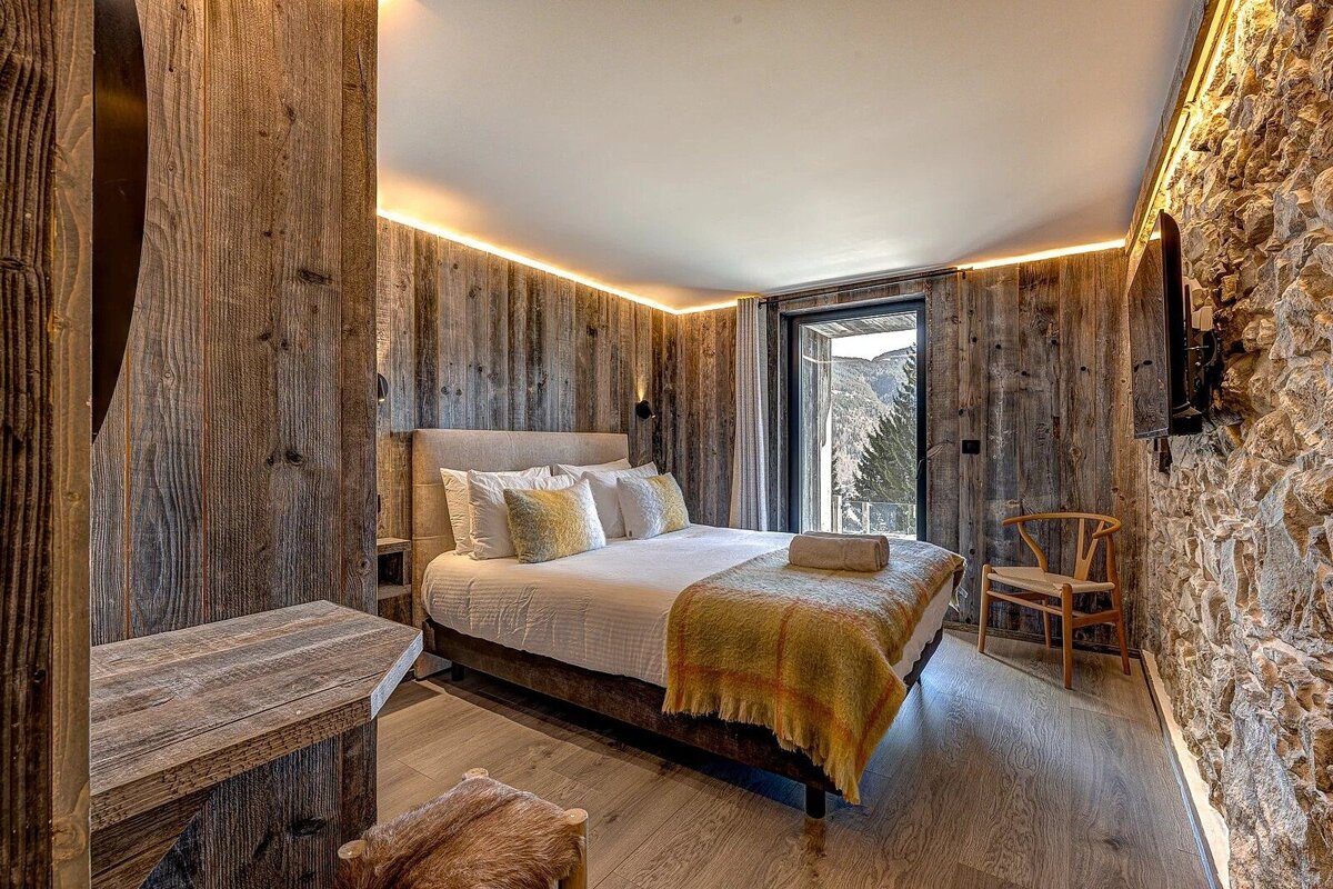 Stylish bedroom with rustic wood & stone walls, modern lighting, a comfy bed, and a dramatic mountain valley view through a large window.