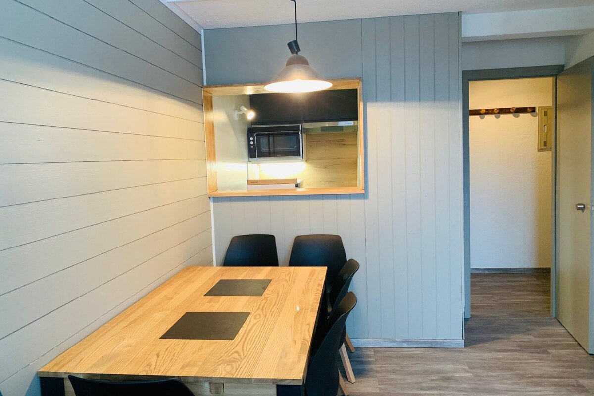 A modern dining area features a wooden table, black chairs, and light grey paneled walls. A kitchen pass-through shows a microwave.