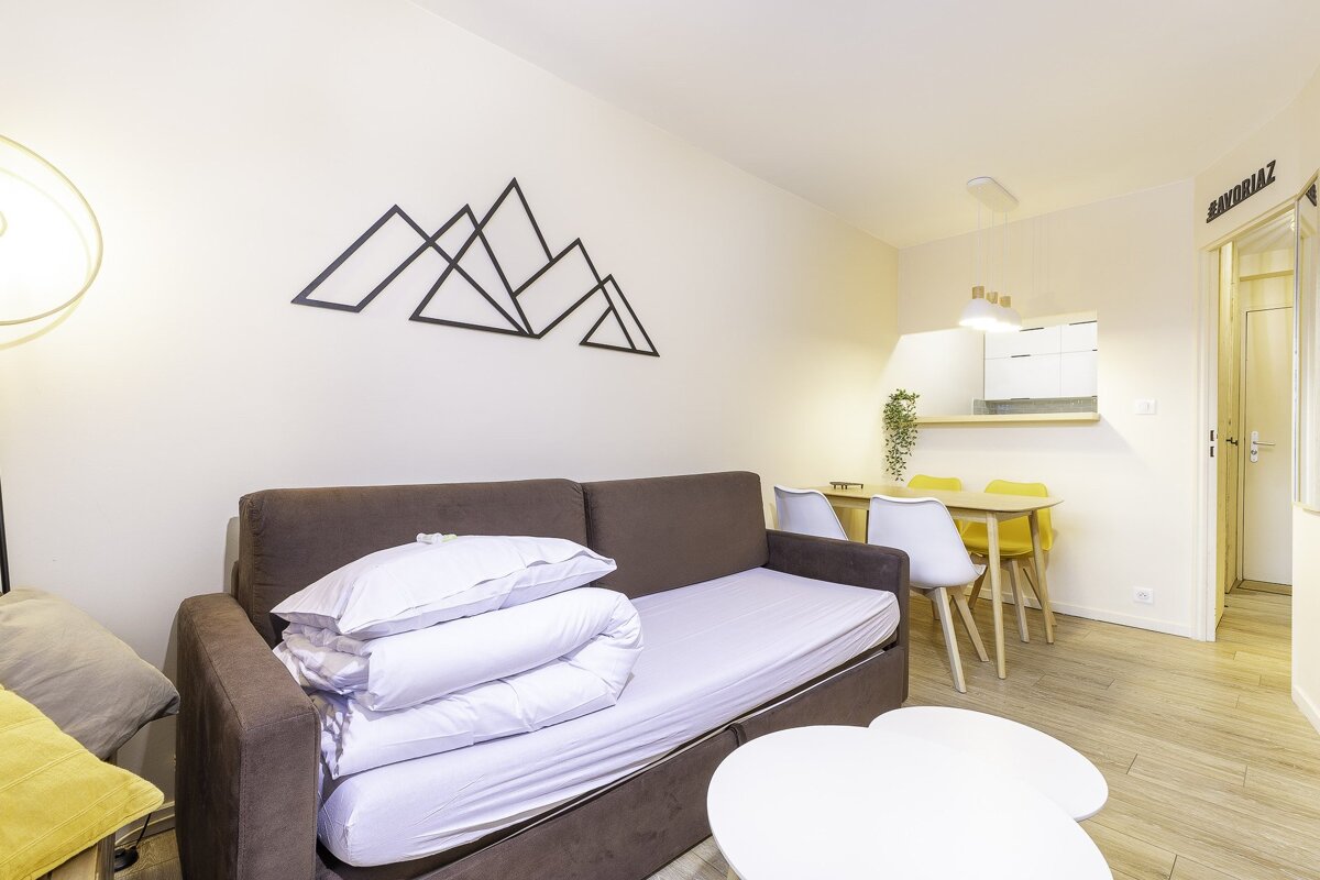 A cozy, modern studio apartment with a brown sofa bed, mountain wall decor, dining area with yellow/white chairs, and a compact kitchenette.