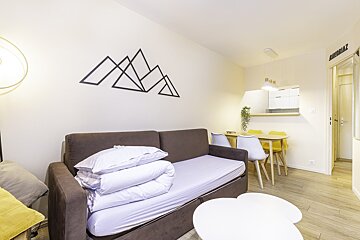 A cozy, modern studio apartment with a brown sofa bed, mountain wall decor, dining area with yellow/white chairs, and a compact kitchenette.