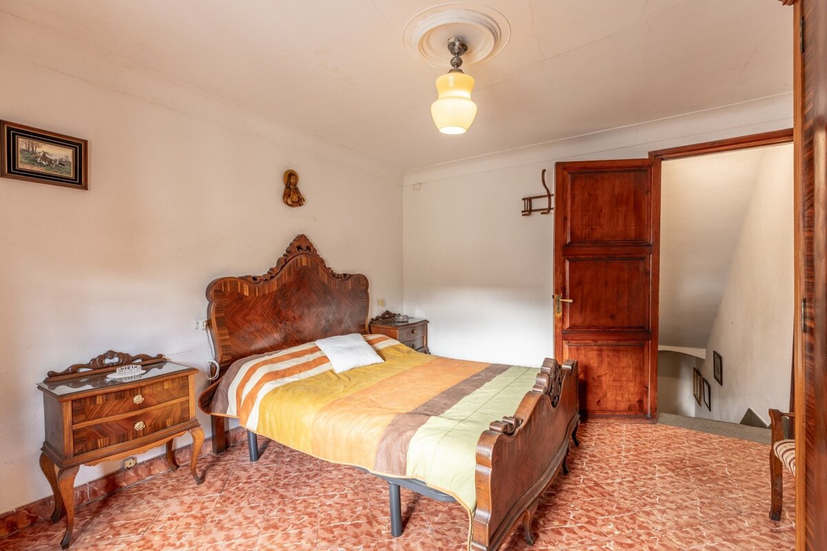Vintage bedroom with ornate wooden bed, striped blanket, and orange tiled floor. An open door leads to stairs. White walls and a retro ceiling lamp.