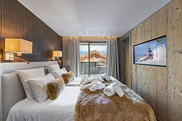 A warm ski chalet bedroom with twin beds, plush brown throw, a TV showing skiing, and a mountain view from the balcony.