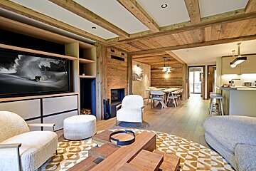 A modern open-plan chalet interior with warm wooden walls, ceiling beams, fireplace, large TV, stylish furniture, dining, and kitchen areas.