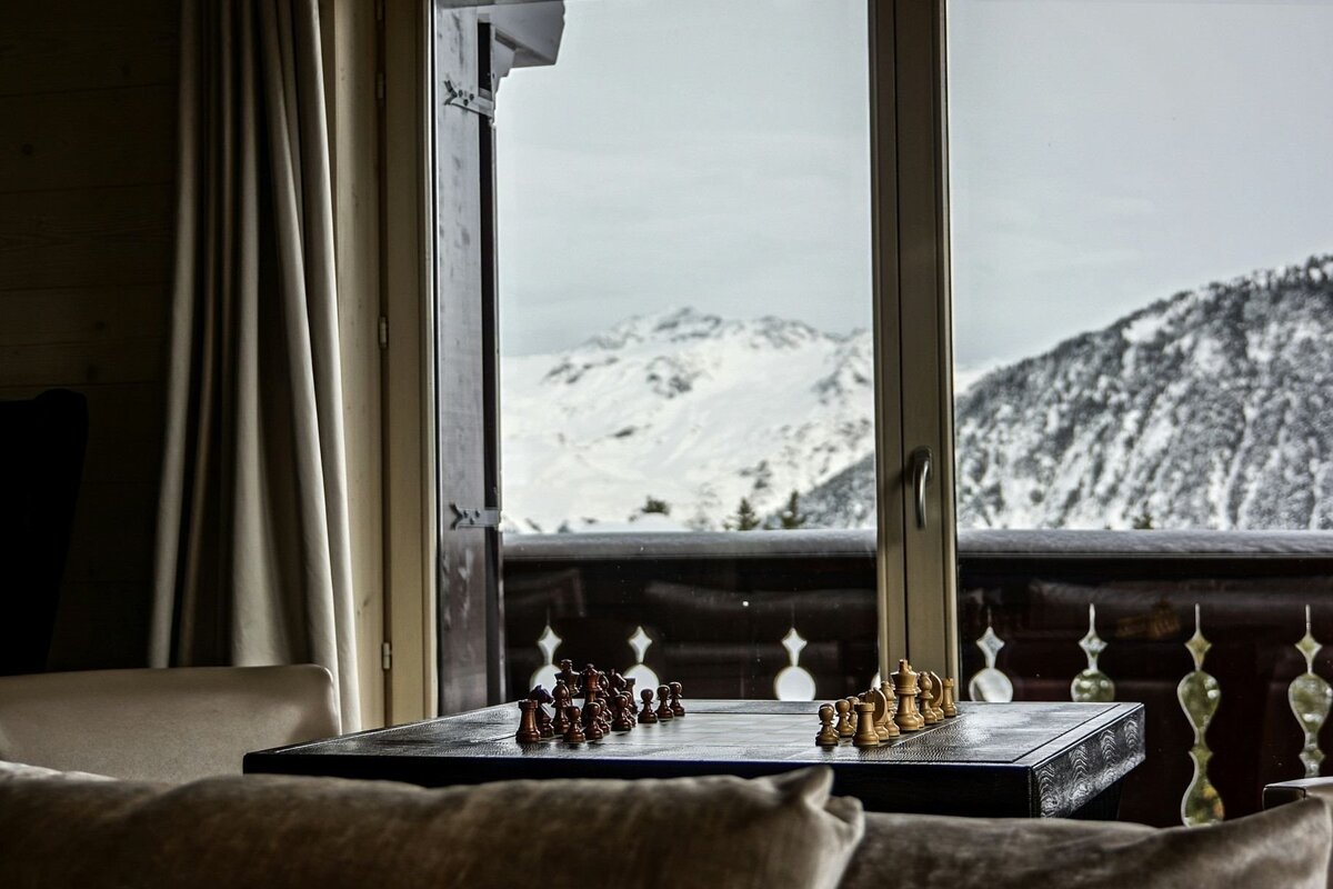 A chess set sits on a table next to a window, framing a breathtaking view of snow-covered mountains and a winter landscape from a cozy room.