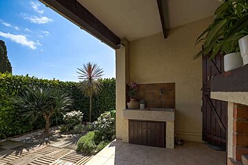 A sunny outdoor patio with an integrated kitchen and a vibrant garden featuring hedges, palm trees, and wooden decking under a clear blue sky.