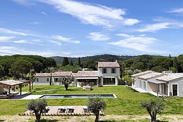 A spacious property with several white houses, red-tiled roofs, a large lawn, swimming pool, and forested hills under a blue sky.