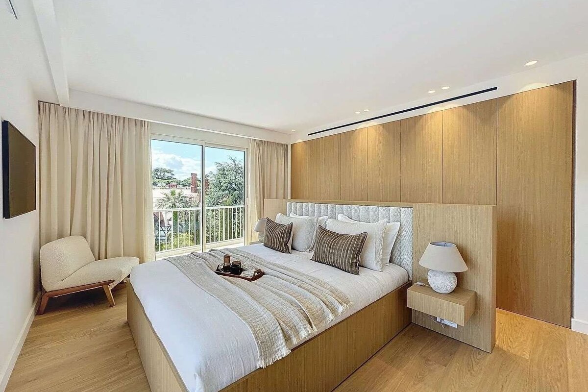 A sleek, modern bedroom features a large bed, light wood paneling, and floor-to-ceiling windows opening to a balcony with greenery. The space is serene and well-lit.