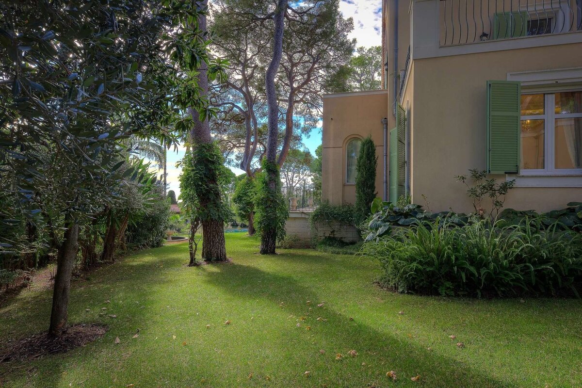 A sunny garden with a green lawn, tall trees, and a beige house with green shutters and a balcony under a bright sky.