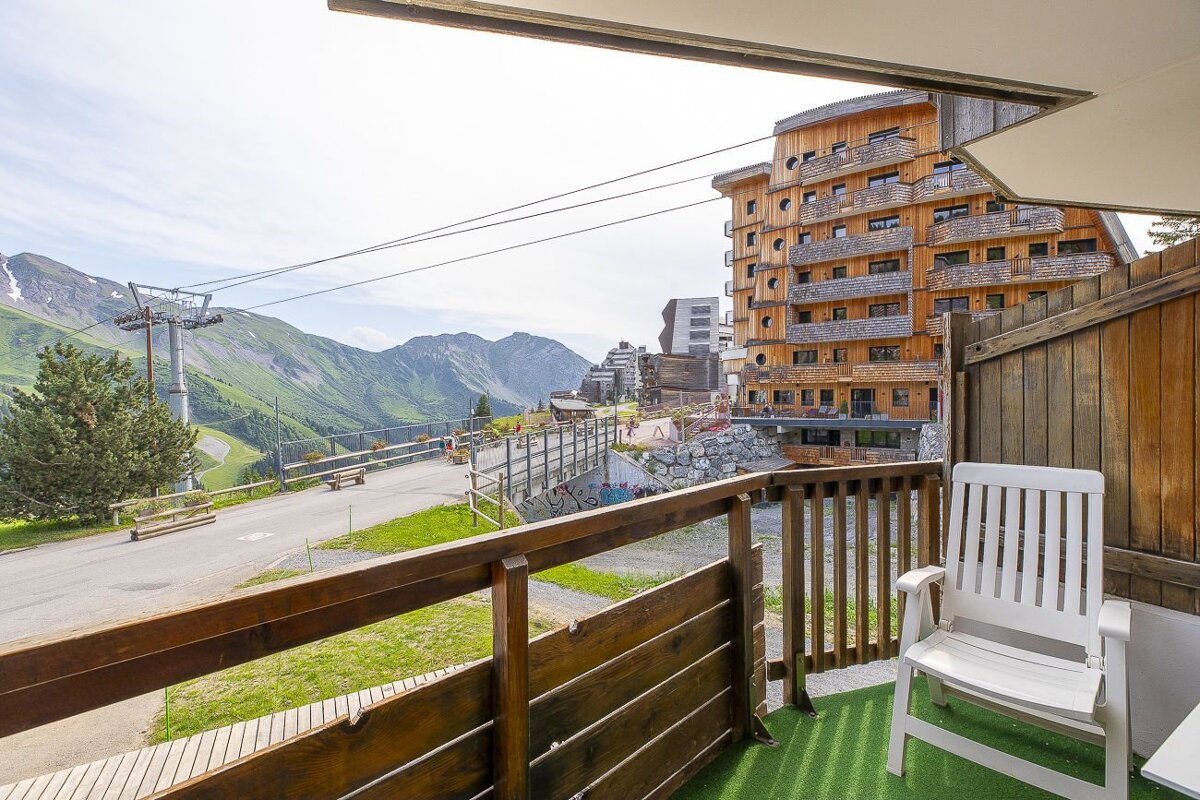 View from a balcony showing a mountain resort with a large wooden building, green slopes, a ski lift, and a road under a bright sky.