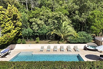 An outdoor pool area with several lounge chairs, a hammock, and a round daybed, set against a backdrop of dense green trees.