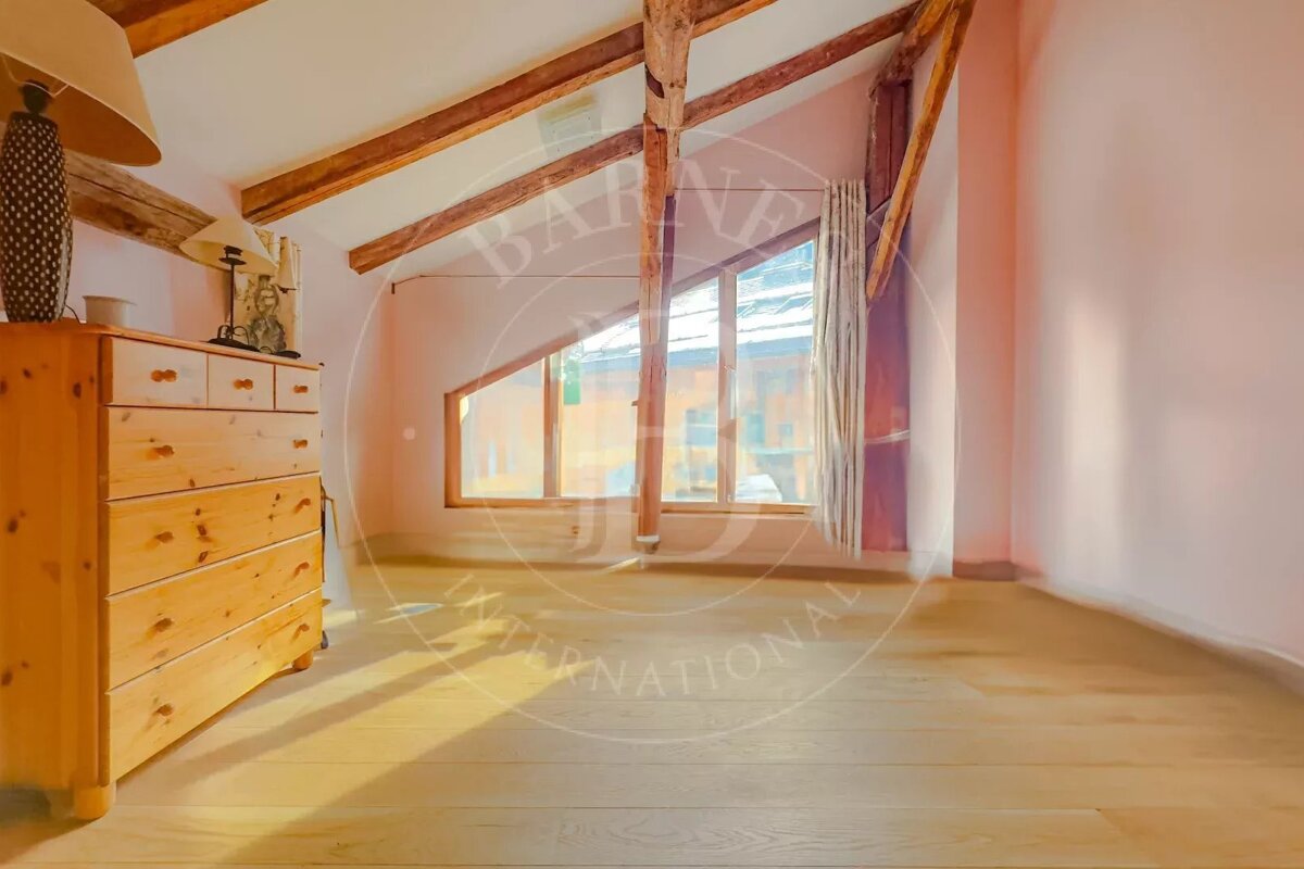 A pink-walled attic room with exposed wooden beams, a slanted window, a wooden dresser with a lamp, and a light wooden floor.