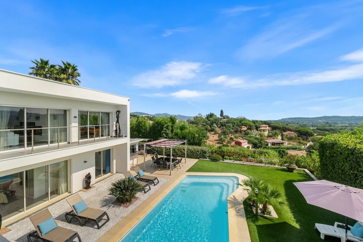 Modern white villa with a swimming pool, backyard lawn, and stunning views of hills under a bright blue sky.
