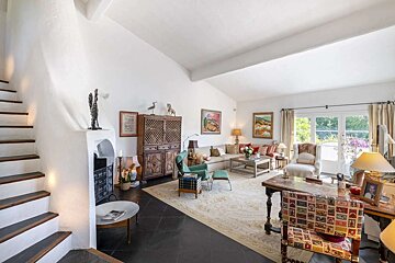 Bright, eclectic living room with white walls, dark floor, staircase, fireplace, various furniture including a sofa, chairs, and large window.
