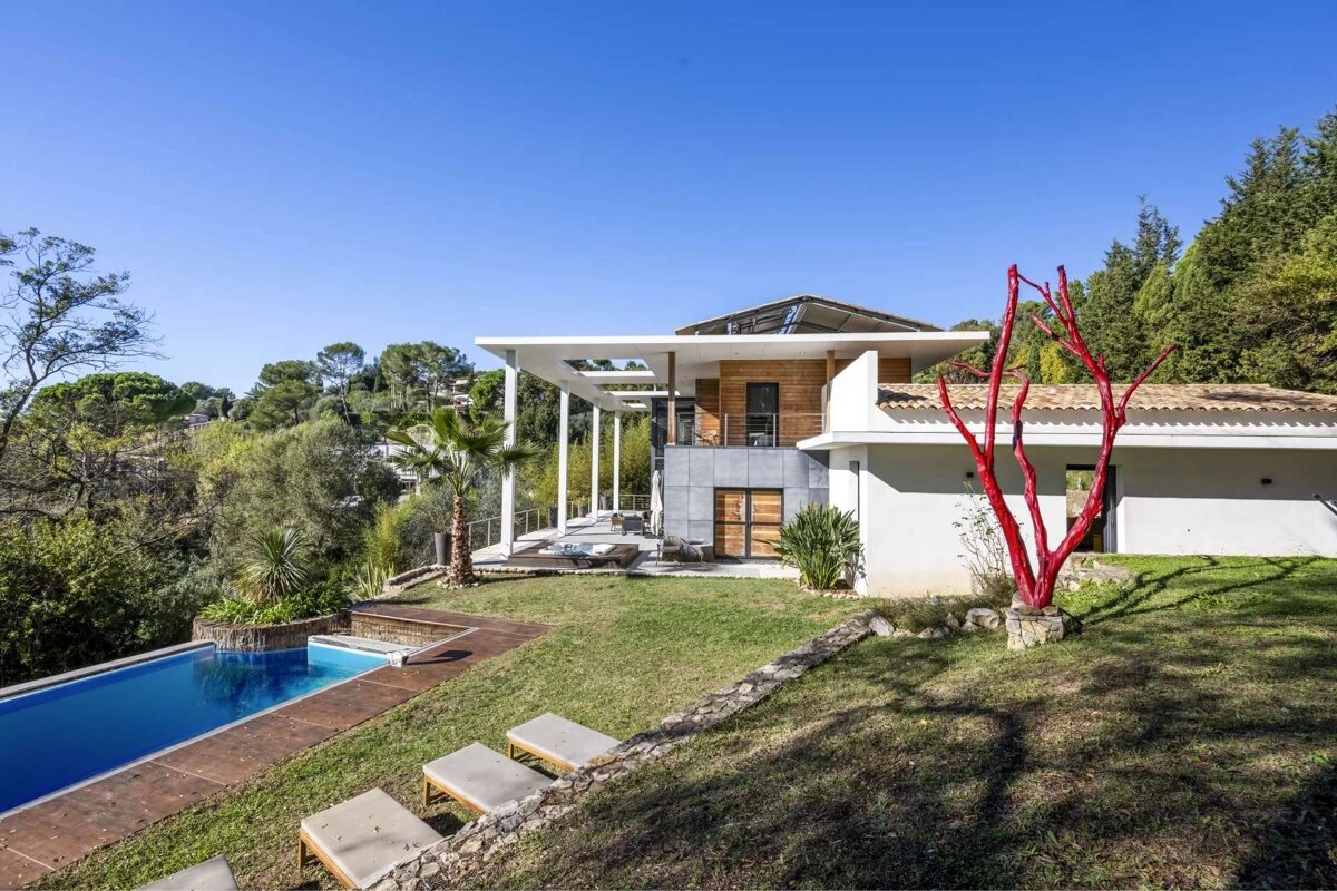 A modern hillside house with an infinity pool and landscaped garden, featuring a striking red tree sculpture under a clear blue sky.
