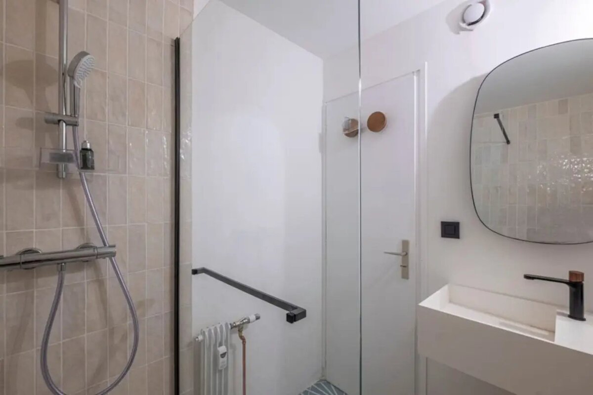 Modern bathroom featuring a beige-tiled shower, glass divider, minimalist white vanity, and a large oval mirror.
