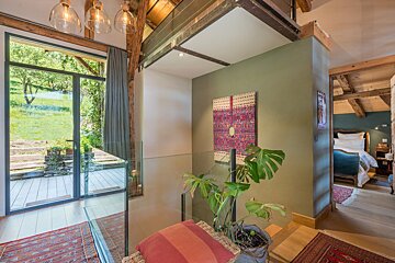 A rustic-modern interior featuring exposed beams, a glass railing, and a view to a lush garden. A cozy bedroom is seen beyond a sage green wall with a tapestry.