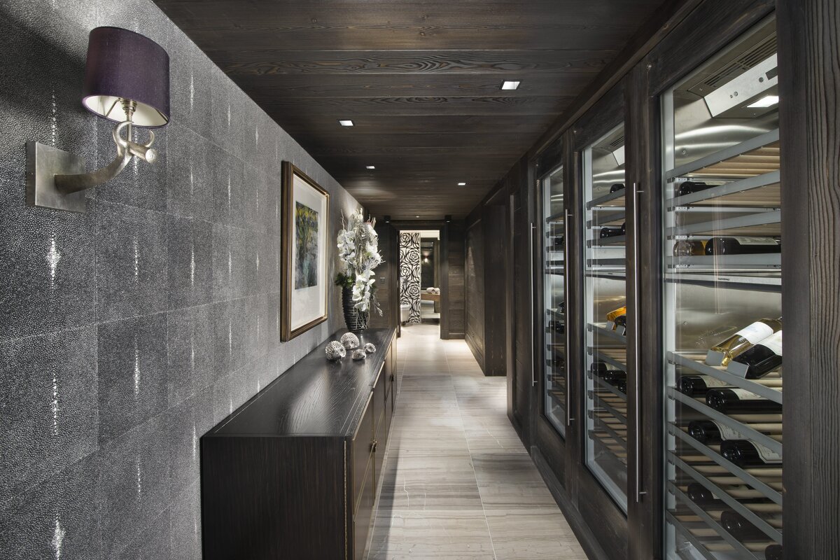 A modern, dark-toned hallway features a wall of wine refrigerators opposite a textured gray wall with a console table and art.