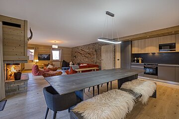 Cozy, open-plan chalet living space with a fireplace, large dining table with fur benches, modern kitchen, and casual seating.