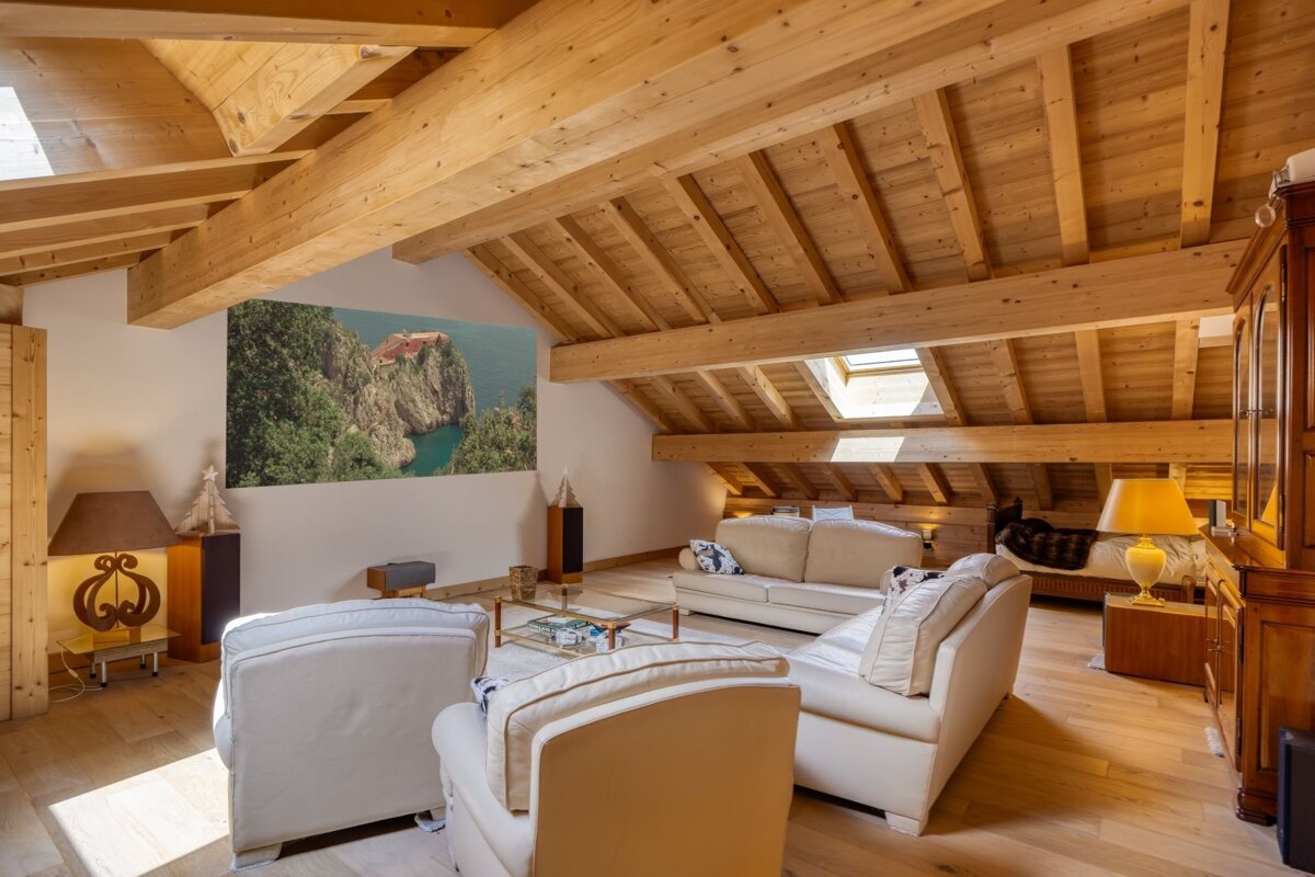 A bright, spacious attic living room features a vaulted wooden ceiling with exposed beams and a skylight. White sofas and armchairs furnish the room, which has a large landscape artwork on one wall.