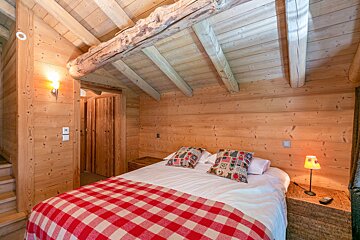 A cozy, rustic wooden bedroom with a bed, red checkered blanket, exposed ceiling beams, and a warm bedside lamp, creating a chalet feel.