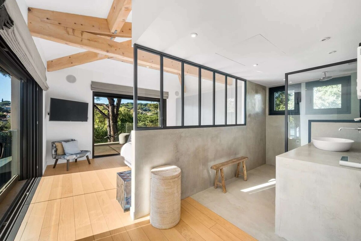 Modern open-plan room with exposed wooden beams, concrete walls, and large windows revealing a lush outdoor view, blending living and bathroom areas.
