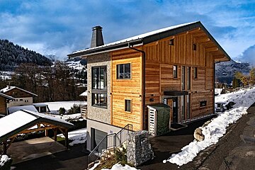 A modern wood and stone house on a snowy slope, with a carport and mountains in the background under a blue sky.