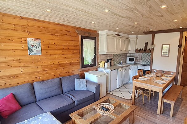 Cozy, rustic open-plan chalet interior with wood-paneled walls & ceiling, featuring a gray sofa, kitchenette, and wooden dining area.
