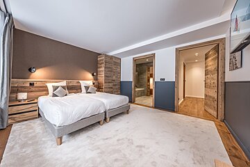Modern bedroom with two twin beds, wooden accents, a white carpet, and two open doorways showing a bathroom and a hallway.