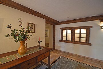A cozy, rustic hallway features exposed wooden beams, a decorated console table, and a faux window with warm light, above a tiled floor.