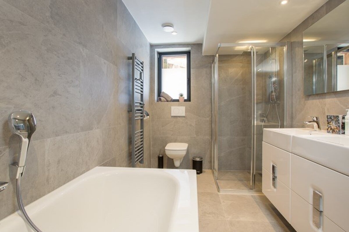 Modern bathroom with grey tiles, featuring a bathtub, glass shower, wall-mounted toilet, and white vanity unit.