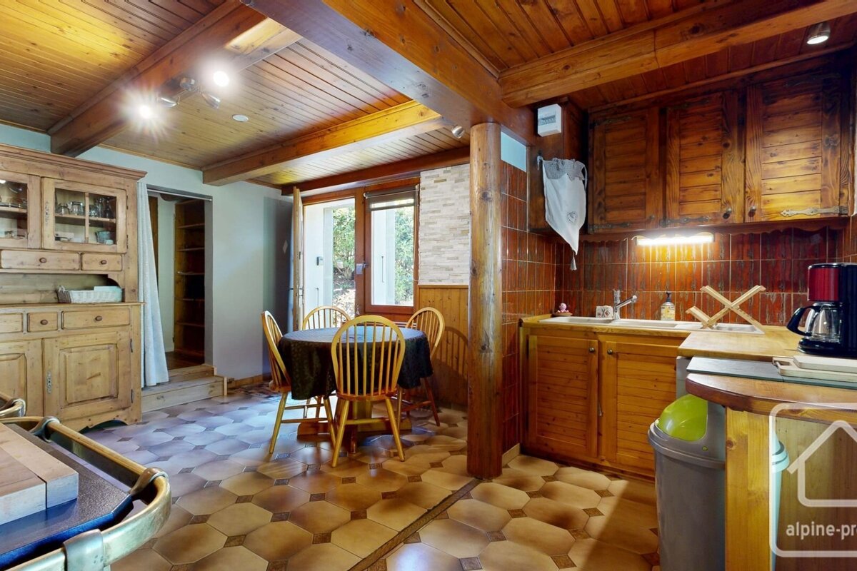 A rustic wooden kitchen and dining area features a patterned tiled floor, exposed beams, solid wood cabinetry, a dining table, and a window.