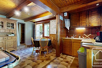 A rustic wooden kitchen and dining area features a patterned tiled floor, exposed beams, solid wood cabinetry, a dining table, and a window.