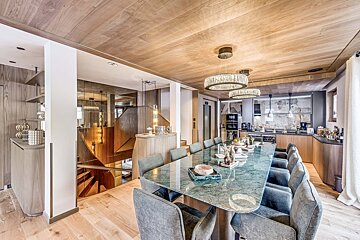 Elegant dining room featuring a green marble table, grey chairs, wooden ceiling, modern chandeliers, and an open staircase.