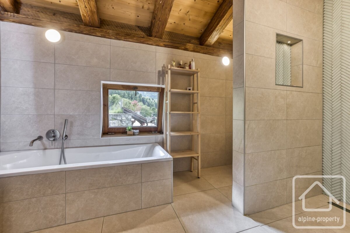 Modern tiled bathroom with a bathtub, wooden ceiling beams, a small window, and a wooden shelf.
