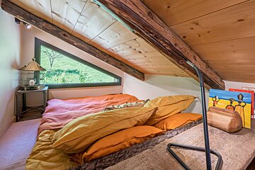 Cozy attic bedroom with a sloped wooden ceiling, rustic beams, colorful bedding, and a unique triangular window framing a lush green view.