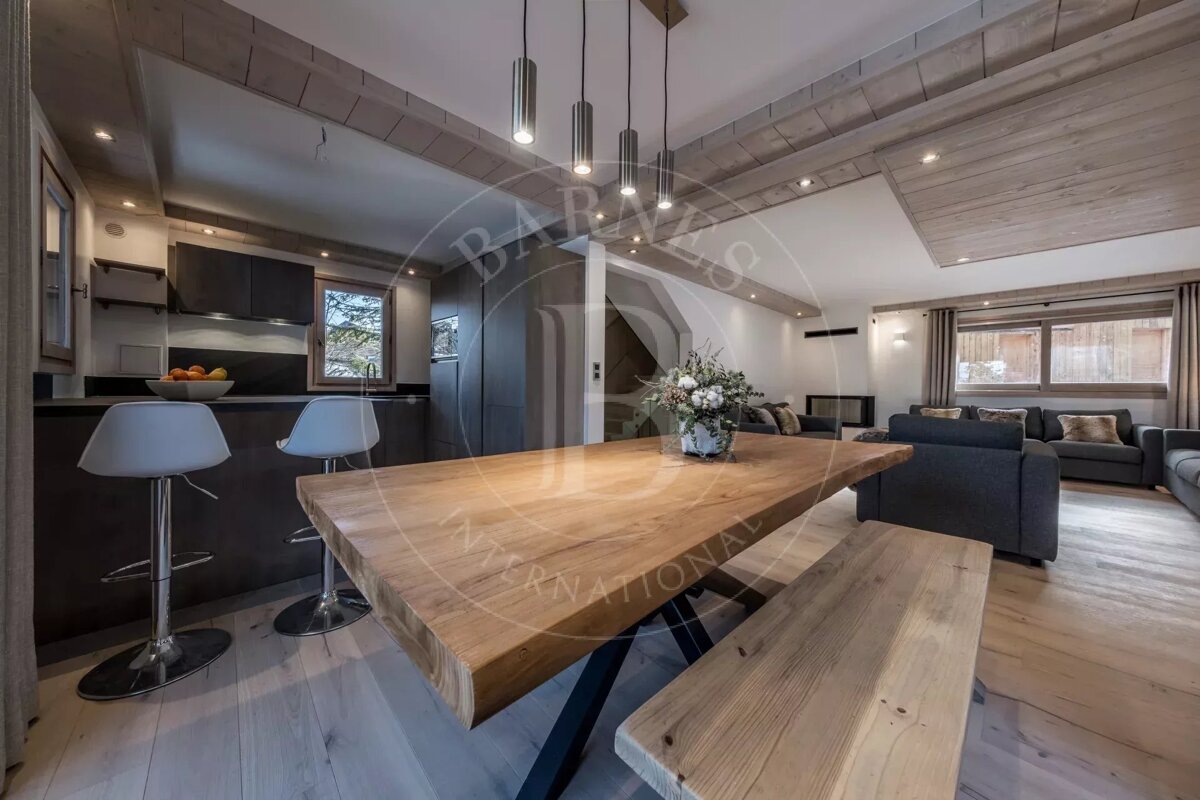 A modern chalet-style open-plan living space featuring a kitchen island, large wooden dining table, and a cozy living room with gray sofas and wood accents.