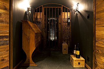 A dark, rustic wine cellar entrance with an ornate iron gate, antique desk, and wine bottles, lit by sconces, leading into a shadowy passage.