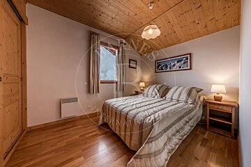 A cozy alpine bedroom with wooden ceiling/floor, a striped bed, warm lighting from lamps, and a window framing a snowy mountain view.
