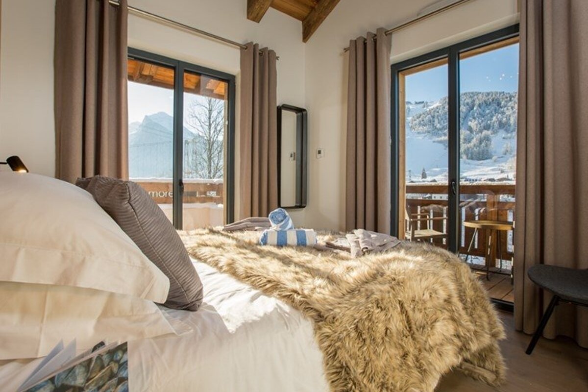A rustic bedroom with a faux-fur blanket, overlooking a snowy alpine mountain landscape through glass balcony doors.